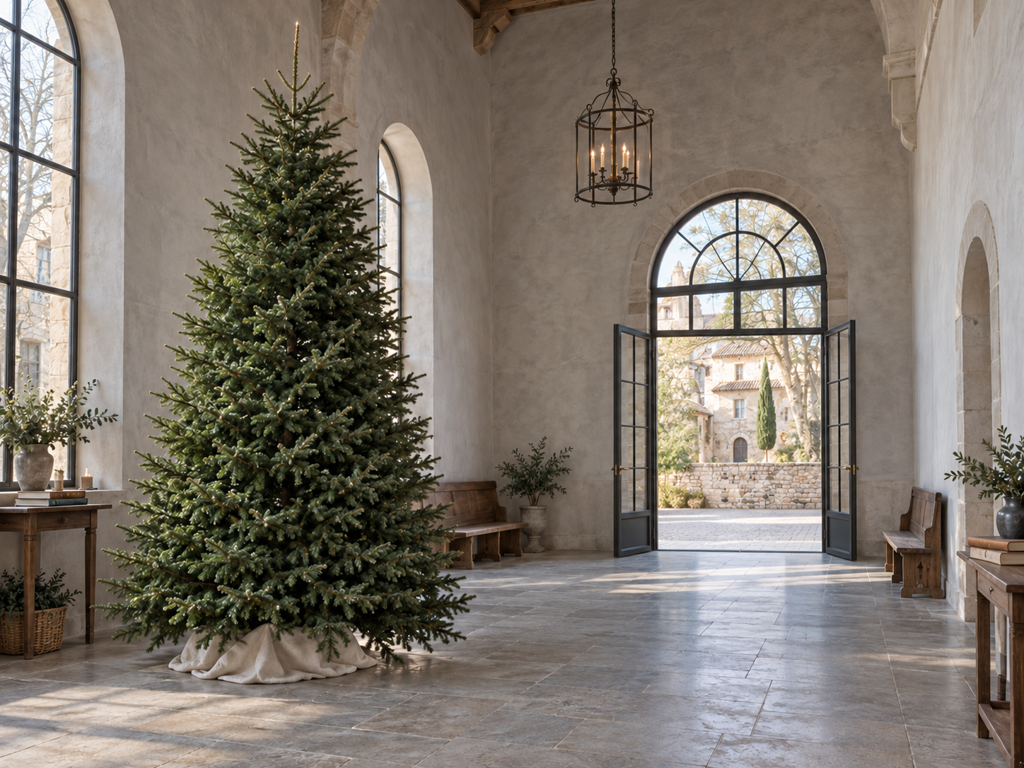 A tall, natural Fraser fir in a peaceful hall with gray plaster walls, aged stone floors, an iron chandelier and open steel French doors.